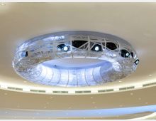 Soffitto interno moderno con grande elemento circolare sospeso, rivestito da una struttura reticolare bianca e dotato di faretti integrati. Intorno si vedono superfici curve illuminate e una fascia di piccole luci incassate nel controsoffitto.