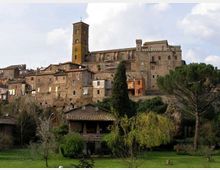 Borgo storico su un’altura con edifici in pietra e una grande chiesa o complesso fortificato con alta torre campanaria. In primo piano si vede un’area verde con alberi e un piccolo edificio con tetto in coppi, sullo sfondo cielo parzialmente nuvoloso.
