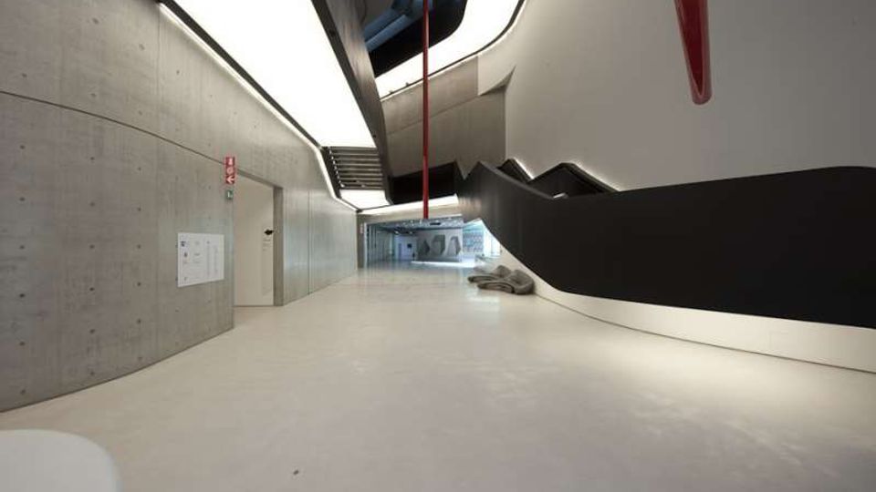 Ampio corridoio interno di un edificio moderno con pavimento chiaro, pareti in cemento a vista e illuminazione lineare a soffitto. Sul lato destro corre una grande struttura/scalinata nera e bianca dalle forme curve, con elementi rossi sospesi e uno spazio aperto visibile in fondo.