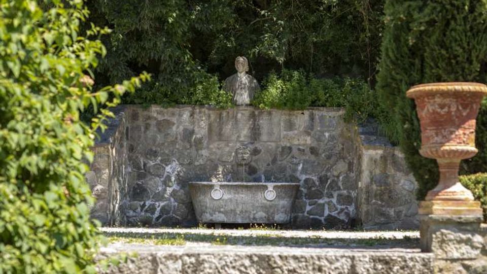 Fontana in pietra in un giardino, con vasca rettangolare e mascherone centrale su un muro rivestito in pietra grezza. Sullo sfondo si vede una statua sopra il muro, circondata da vegetazione fitta, e a destra un grande vaso ornamentale in terracotta su piedistallo.