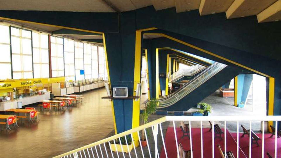 Ampio atrio interno di un edificio moderno con grandi finestre a tutta altezza sul lato sinistro, tavoli e sedie disposti come area ristoro. Al centro si vedono travi e pilastri angolari blu e gialli e una serie di scale mobili che attraversano lo spazio.