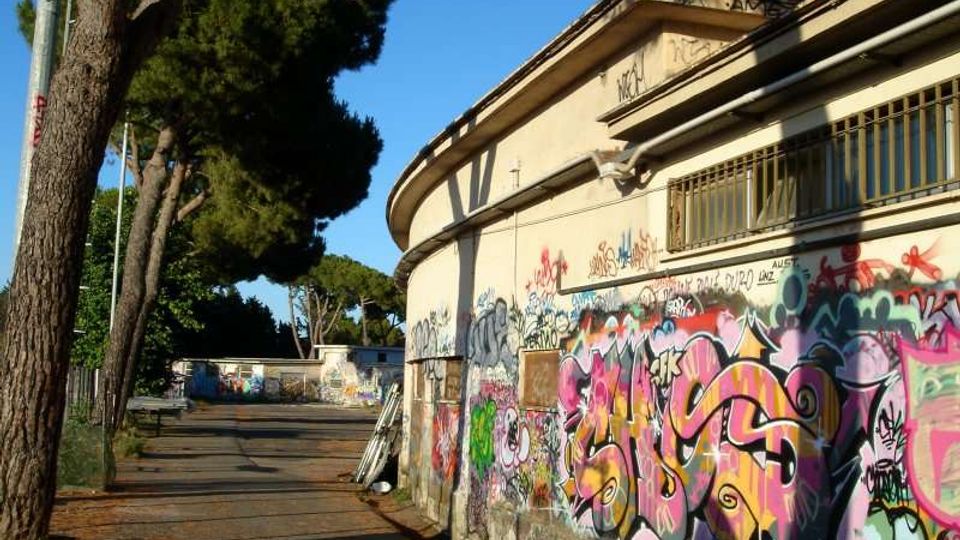 Edificio basso con facciata curva e finestre con grate, ricoperto di graffiti colorati lungo un vialetto asfaltato. A sinistra si vedono alti pini e un’area esterna con altri muri dipinti, sotto un cielo sereno.