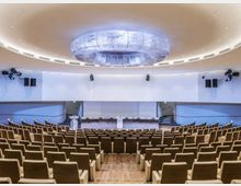Auditorium moderno con file di poltrone in legno e tessuto disposte a gradoni, rivolte verso un palco con grande schermo bianco e podio centrale. Soffitto curvo con ampia struttura circolare luminosa e impianti audio e luci laterali.