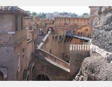 Vista di un complesso fortificato in mattoni con mura merlate, archi e passaggi sopraelevati. Un ponte pedonale metallico collega le strutture, con tetti in tegole e pareti in pietra visibili ai lati.