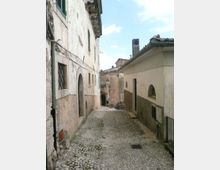 Vicolo stretto in pietra con pavimentazione a ciottoli, fiancheggiato da vecchi edifici in intonaco con porte ad arco e finestre con persiane. La strada in lieve discesa conduce verso altre case addossate, sotto un cielo parzialmente nuvoloso.