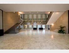 Ampio atrio interno di un edificio moderno con pavimento in marmo chiaro, pareti in pietra e grandi finestre sul fondo. A sinistra c’è una scala in pietra con corrimano, mentre a destra si vede un’area con piante e una panca, illuminata da luci a parete e a soffitto.