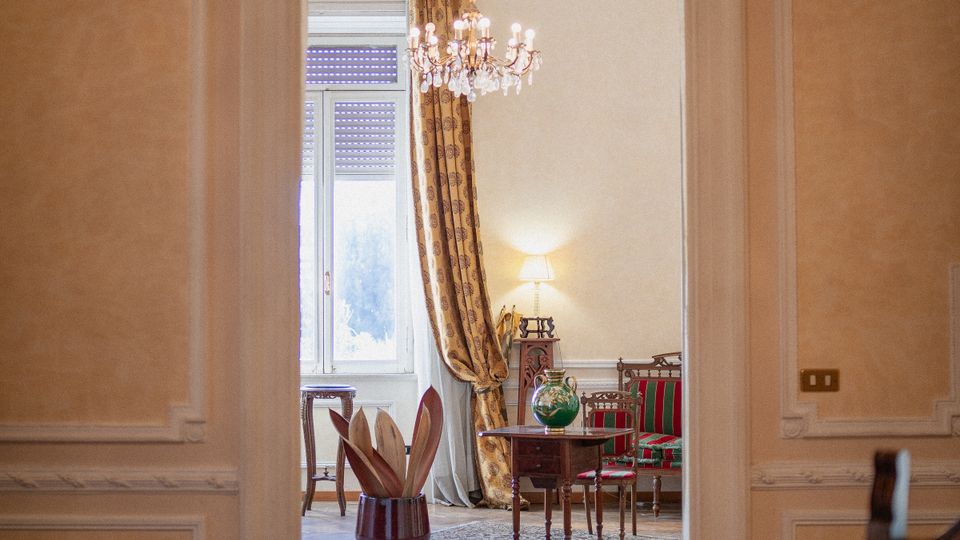 Interno di un appartamento elegante con pareti in stucco e cornici decorative, con un arco sopra la porta che incornicia un dipinto ovale di fiori. Attraverso la porta si vede un salotto con lampadario, grandi finestre con tende pesanti, parquet a spina e mobili classici con poltrone a righe e tavolino.