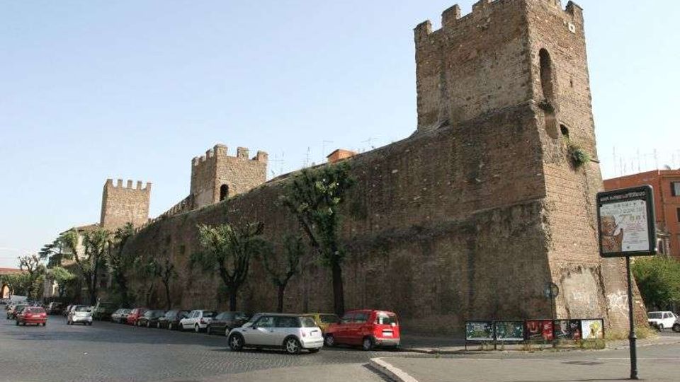 Lungo tratto di mura medievali in mattoni con torri merlate, affacciato su una strada cittadina con auto parcheggiate e alberi allineati. Sullo sfondo si vedono edifici urbani e un cartello stradale vicino al marciapiede.