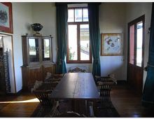 Sala da pranzo con lungo tavolo in legno e sedie intagliate, pavimento in legno e credenza vetrata. Una grande finestra con tende verde scuro illumina l’ambiente, con quadri alle pareti e porta finestrata sul lato destro.