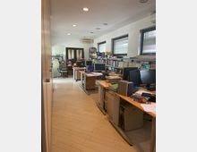Interno di un ufficio condiviso con più scrivanie in fila, computer e pile di documenti, affiancato da scaffali pieni di raccoglitori e libri. Pareti bianche con finestre dotate di veneziane, pavimento in piastrelle chiare e illuminazione a faretti incassati nel soffitto.