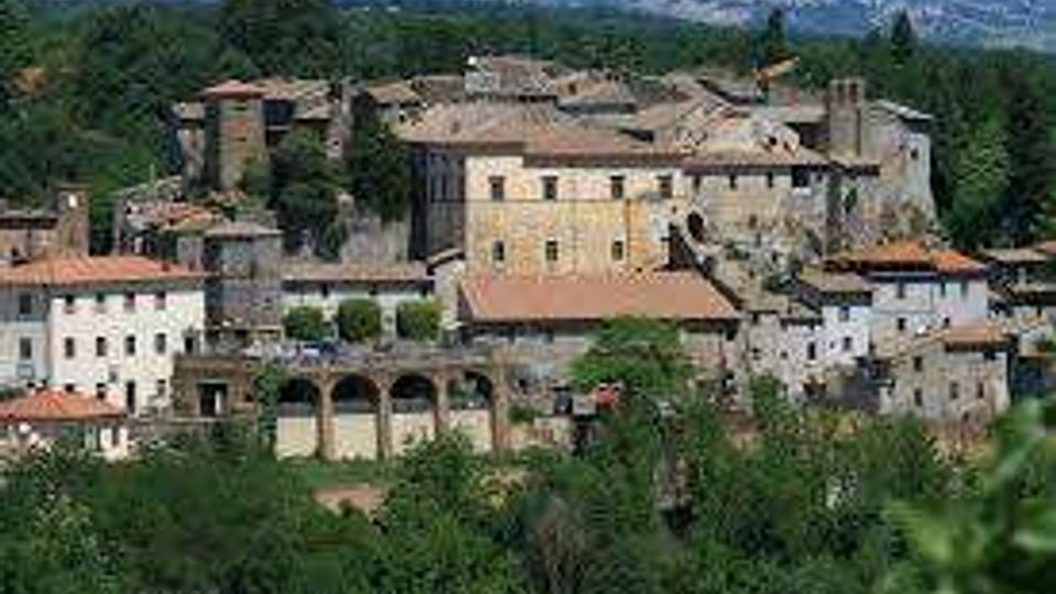 Veduta panoramica di un borgo storico su una collina, con edifici in pietra dai tetti in tegole e una struttura ad archi nella parte inferiore. Il paese è circondato da colline verdi e vegetazione, con l’orizzonte della campagna sullo sfondo.