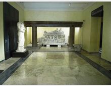 Sala museale con pavimento in marmo e pareti chiare, con al centro una struttura in pietra a colonne che incornicia un grande sarcofago scolpito in rilievo. A sinistra è esposta una statua su piedistallo e a destra si vede l’ingresso verso un’altra area espositiva.