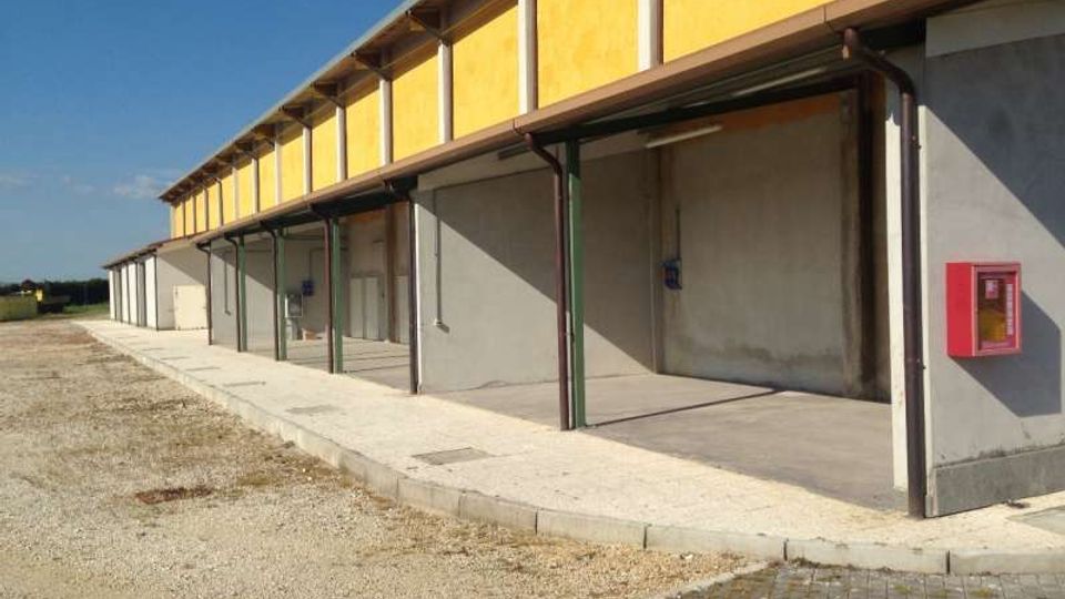 Lungo edificio industriale a un piano con fascia superiore gialla e pareti grigie, dotato di tettoia sostenuta da pilastri metallici lungo una banchina in cemento. Davanti si vede un ampio piazzale in ghiaia e pavimentazione, con un armadietto antincendio rosso sulla parete laterale.