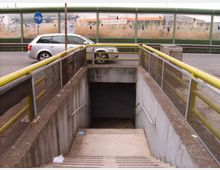 Ingresso a un sottopassaggio pedonale con scala in cemento che scende sotto la strada, affiancata da corrimani e parapetti con ringhiere gialle. Sullo sfondo si vede la carreggiata con un’auto e una barriera fonoassorbente verde lungo la strada.