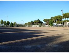 Ampio campo sportivo in terra battuta recintato, con un cancello e pali dell’illuminazione; sullo sfondo si vedono alberi ad alto fusto e alcune strutture basse sotto un cielo sereno.
