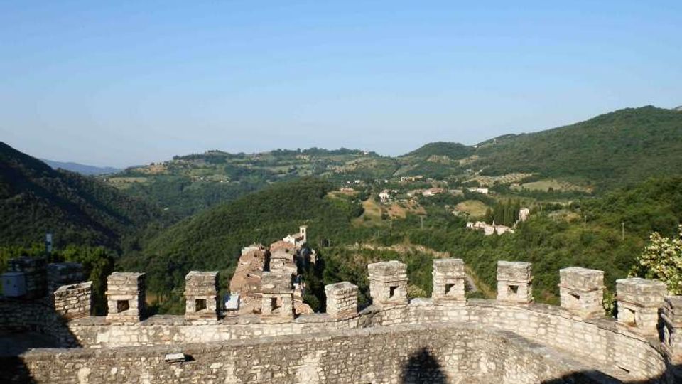 Vista panoramica da una terrazza o camminamento di un castello in pietra, con merlature quadrate in primo piano. Sullo sfondo si vedono colline e vallate verdi con piccoli borghi e campi sotto un cielo sereno.