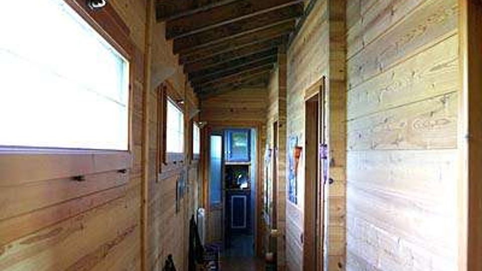 Corridoio interno stretto con pareti e soffitto in legno a vista, travi scure inclinate e pavimento piastrellato. Sul lato sinistro ci sono finestre che illuminano l’ambiente, mentre a destra si aprono porte lungo il corridoio fino a una porta in fondo.