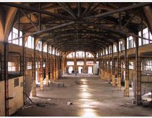 Interno di un grande capannone industriale abbandonato, con ampia navata centrale e struttura a capriate metalliche a vista sotto il tetto. Lungo le pareti si vedono finestre alte e pilastri in muratura, con pavimento in cemento parzialmente coperto da detriti e aperture sul fondo.