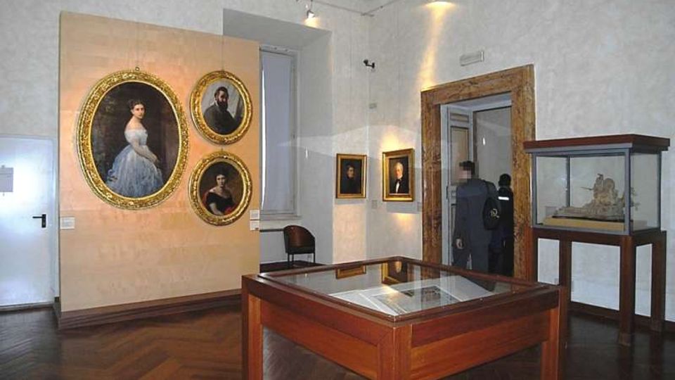 Sala museale con pareti chiare e pavimento in parquet, con grandi ritratti incorniciati appesi alle pareti e teche espositive in legno e vetro al centro e lungo il lato destro. Un’ampia apertura con cornice in legno conduce a un’altra stanza, illuminata da faretti a soffitto.