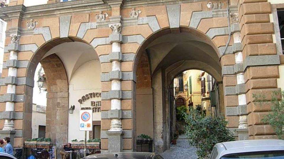 Facciata di un edificio in stile storico con grandi arcate in pietra e muratura bugnata, che formano un portico con volte e un passaggio pedonale verso una stretta via interna. Davanti alle arcate si vedono auto parcheggiate e, sotto il portico, arredi e insegne di un locale.