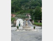 Piazza di un piccolo paese con una fontana in pietra al centro, su basamento circolare con catene e vasi. Sullo sfondo si vede una cappella bianca con porta verde, una strada che sale tra case sparse e un pendio collinare coperto di alberi.
