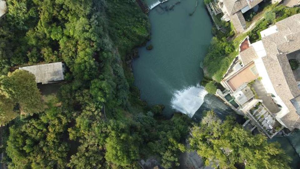 Vista aerea di un piccolo fiume con una diga o sbarramento che crea una breve cascata, circondato da vegetazione fitta. A lato si vedono edifici con tetti in tegole e una struttura rettangolare in muratura vicino al bosco.