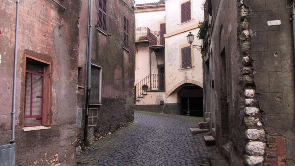 Vicolo stretto in un centro storico con pavimentazione in sampietrini, fiancheggiato da edifici in muratura intonacata e pietra con finestre a persiane e tubazioni a vista. In fondo si vede una casa chiara con balconcino, lampione a muro e un arco che conduce a un passaggio coperto.