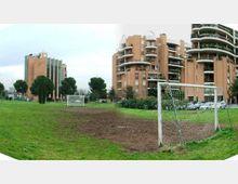 Campo da calcio in erba con aree di terra battuta e due porte, situato in un parco urbano. Sullo sfondo si vedono alti edifici residenziali in mattoni con balconi curvi, alberi e auto parcheggiate.
