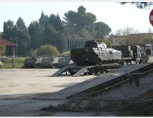 Piazzale asfaltato in un’area militare con un carro armato su un rimorchio a pianale ribassato collegato a una rampa metallica. Sullo sfondo si vedono altri mezzi corazzati in fila, edifici bassi e alberi lungo il perimetro.
