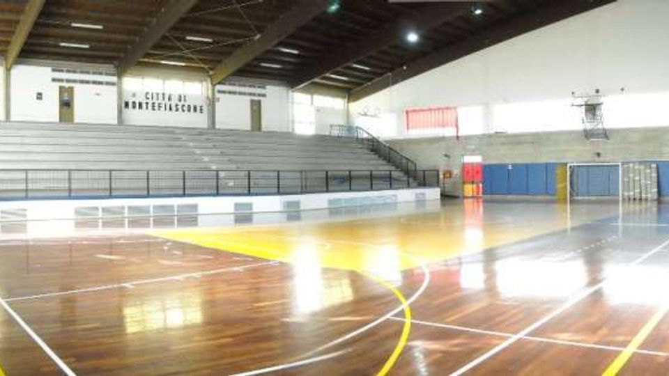 Interno di un palazzetto dello sport con campo polivalente in parquet lucido e linee tracciate per diversi sport. Sul lato lungo si vedono ampie gradinate, pareti con pannelli colorati e un soffitto alto con travi e illuminazione.