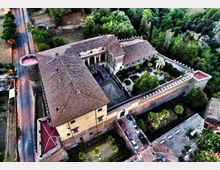 Veduta aerea di un grande complesso in muratura con tetti in coppi, organizzato attorno a un cortile interno e circondato da mura merlate con torri agli angoli. All’interno delle mura si vede un giardino alberato e, all’esterno, una strada che corre lungo il lato sinistro dell’edificio.