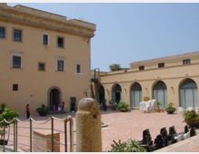 Cortile lastricato in mattoni tra edifici color crema, con un alto palazzo a più piani e un porticato ad archi con grandi vetrate sul lato destro. In primo piano si vede un pilastro in pietra e, lungo il perimetro, catene tra paletti, piante in vaso e alcune sedie impilate.