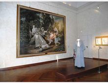 Sala museale con pavimento in parquet e pareti bianche, dominata da un grande dipinto incorniciato appeso alla parete. Al centro della stanza è esposto su una pedana un abito storico, con faretti e alcune opere incorniciate visibili sulla parete laterale.