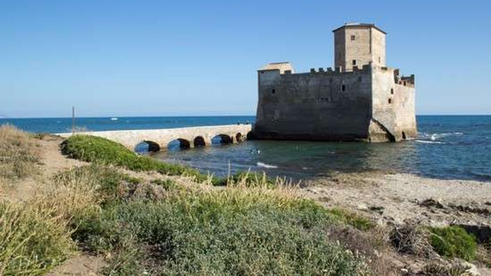 Fortezza in pietra sul mare con torre quadrata e mura merlate, collegata alla riva da un ponte in muratura con archi. In primo piano si vede una costa rocciosa con vegetazione bassa e, sullo sfondo, il mare aperto sotto un cielo sereno.