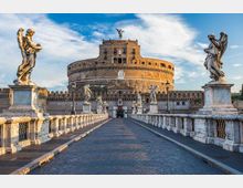 Veduta frontale del Castel Sant’Angelo con la sua struttura circolare in pietra, visto dal ponte con pavimentazione in selciato e balaustre in marmo. Lungo il ponte sono presenti statue di angeli su piedistalli, con cielo azzurro e nuvole sullo sfondo.