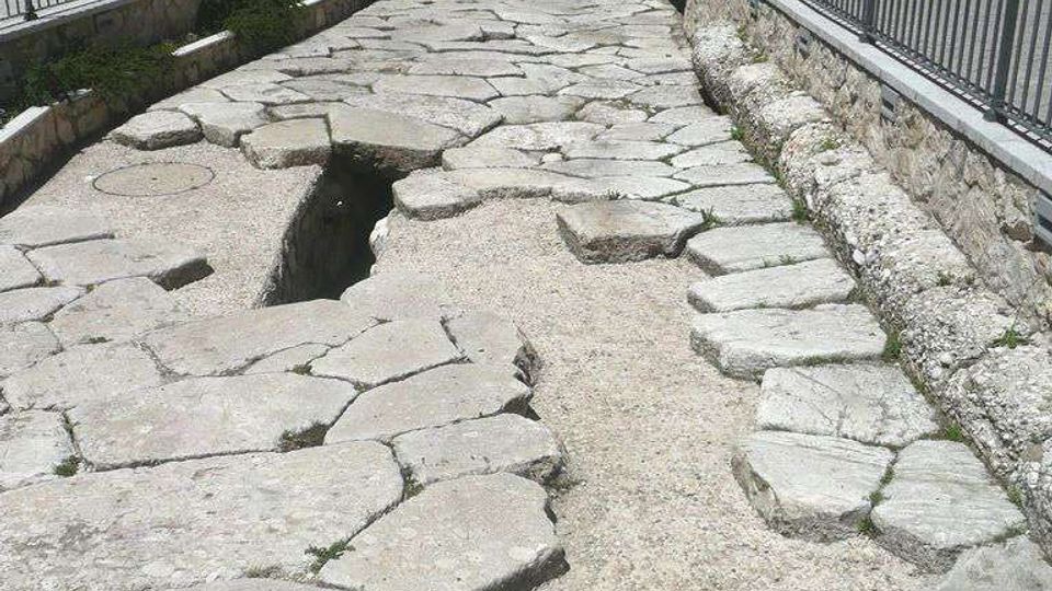 Antica strada lastricata in grandi pietre irregolari, incassata in una trincea e delimitata da muretti in pietra e ringhiere metalliche. Sullo sfondo si vede una piazza con auto parcheggiate e un edificio storico con archi e targhe sulla facciata.