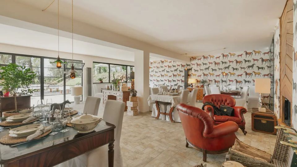 Ampio soggiorno-sala da pranzo open space con pavimento in piastrelle chiare, grandi vetrate scorrevoli e parete decorata con carta da parati a motivo di cavalli. Arredi misti includono tavoli apparecchiati con sedie rivestite, poltrone in pelle bordeaux, lampade a sospensione e mobili in legno.