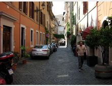 Strada stretta in pavé con auto parcheggiate ai lati, fiancheggiata da palazzi alti con facciate colorate, persiane e lampioni. Vasi e grandi fioriere con alberelli costeggiano i marciapiedi e la via prosegue in prospettiva verso un edificio sullo sfondo.