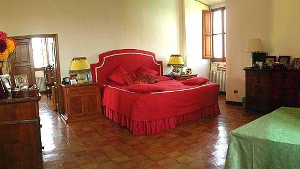 Ampia camera da letto con pavimento in cotto, letto matrimoniale con testiera rossa al centro e un secondo letto singolo con copriletto verde in primo piano. La stanza è arredata con comodini, cassettiere e specchiera, e illuminata da una finestra con infissi in legno.
