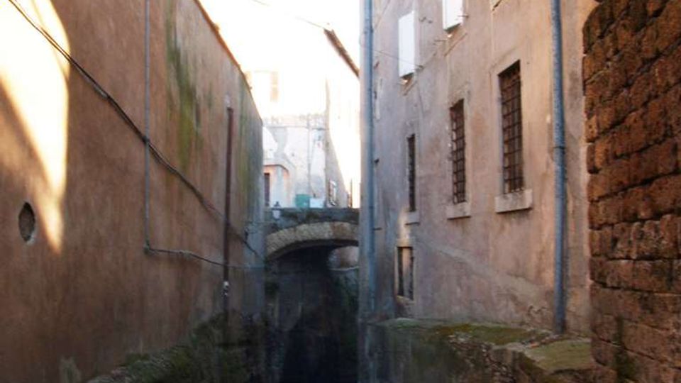 Vicolo stretto in pendenza tra alti edifici in muratura intonacata, con finestre sbarrate e tubazioni esterne lungo le pareti. In fondo si vede un piccolo ponte ad arco in pietra sopra la strada, con muschio e segni di umidità sulle basi dei muri.