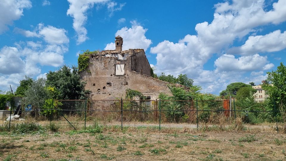 Edificio rurale in rovina con mura scrostate e tetto parzialmente crollato, con un piccolo campaniletto sulla sommità. Davanti c’è una recinzione metallica e vegetazione incolta, con alberi e alcune case sullo sfondo sotto un cielo azzurro con nuvole.