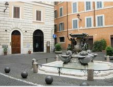 Piccola piazza lastricata con una fontana in pietra al centro, decorata con figure scolpite e vasche circolari con acqua. Intorno si vedono palazzi storici intonacati con finestre e portoni ad arco, lampioni a parete e dissuasori sferici lungo il bordo della piazza.