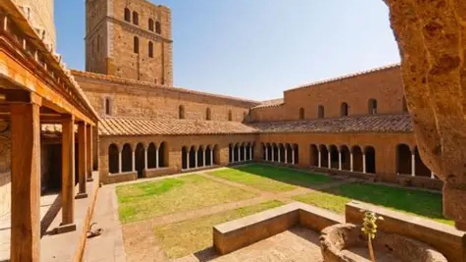 Chiostro di un complesso monastico in pietra con portici ad arcate su tre lati, cortile centrale erboso e un antico pozzo in primo piano. Sullo sfondo si vede una torre campanaria quadrata e edifici con tetti in tegole sotto un cielo sereno.
