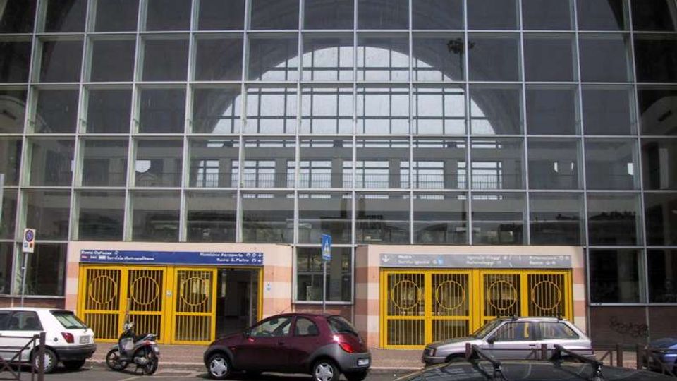 Facciata di una grande stazione ferroviaria con ampia vetrata a griglia e arco superiore, che lascia intravedere la struttura interna. Alla base ci sono ingressi con cancellate gialle e insegne, affacciati su una strada con auto parcheggiate.
