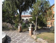 Piccolo parco urbano con vialetto in pietra decorato a motivi circolari bianchi e due pilastri in mattoni con vasi all’ingresso. L’area è circondata da alberi e aiuole, con edifici storici sullo sfondo e una strada laterale.