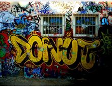 Muro esterno di un edificio ricoperto da graffiti multicolori, con una grande scritta gialla “DONUT” in primo piano. In alto si vedono due finestre rettangolari con inferriate metalliche.
