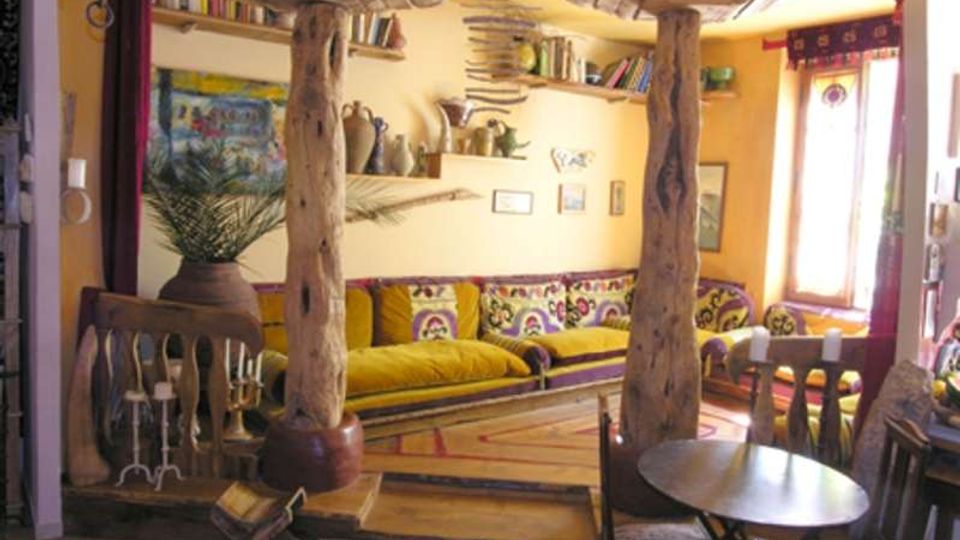 Interno di un soggiorno rustico con travi e colonne in legno grezzo, divano lungo con cuscini decorati e mensole a parete con libri e oggetti. In primo piano si vedono un piccolo tavolo con sedie e, sullo sfondo, una finestra che illumina la stanza.