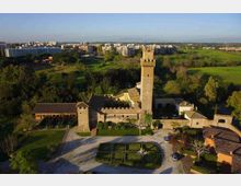 Veduta aerea di un complesso storico con torre in mattoni e mura merlate, circondato da un giardino geometrico e vialetti. Sullo sfondo si estendono campi verdi e un’area urbana con palazzi residenziali.