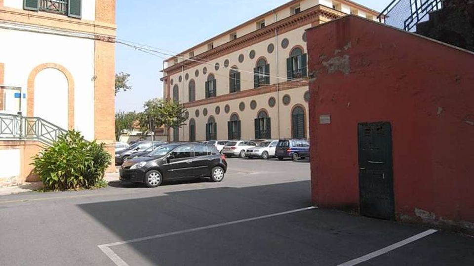 Parcheggio asfaltato con auto in sosta davanti a un edificio storico a due piani con facciata chiara, finestre ad arco e decorazioni circolari. In primo piano si vede un muro rosso con una porta e, a sinistra, parte di un edificio con persiane verdi.