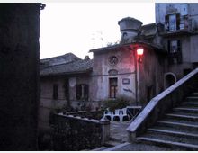 Cortile in un borgo storico con edifici in pietra intonacata e tetti in tegole, illuminato da un lampione rosso vicino a una piccola facciata con finestre tonde. In primo piano si vedono una scalinata in pietra con parapetto e alcune sedie e un tavolo in plastica su una terrazza.